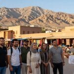 Tour e Pacchetti per il Marocco: Guida Completa 2026 2 A stunning Moroccan landscape featuring an exclusive tour scene. In the foreground, a diverse group of travelers in modest casual clothing, happily engaging with a local guide, showcasing authentic cultural experiences. In the middle ground, traditional Moroccan architecture, such as vibrant riads and market stalls filled with local goods. The background features the Atlas Mountains bathed in natural light under a clear blue sky, enhancing the picturesque setting. Soft, warm lighting creates a welcoming and adventurous atmosphere, capturing the essence of unique Moroccan holidays. The image should convey excitement and authenticity, inviting viewers to explore the wonders of Morocco. Persone che visitano il mercato marocchino, paesaggio suggestivo.